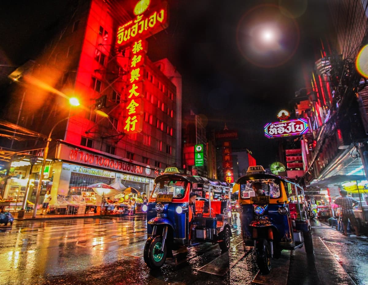 Tuk-tuks parked on a neon-lit street in Bangkok's Chinatown at night, wet from rain